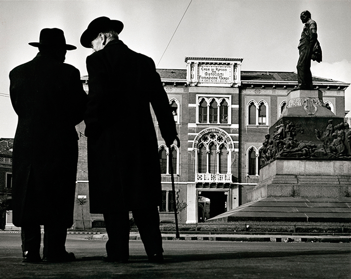 Men in dark trenchcoats with hats, one with cane, next to bronze statue